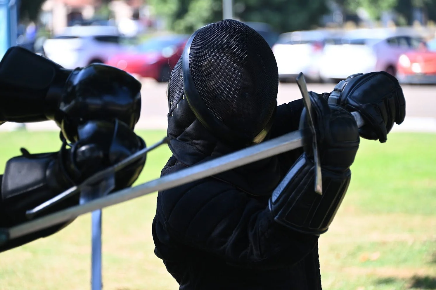 A person dressed in black fencing gear, including a mesh mask and gloves, is practicing fencing outdoors with a sword, against a background of parked cars and green grass.