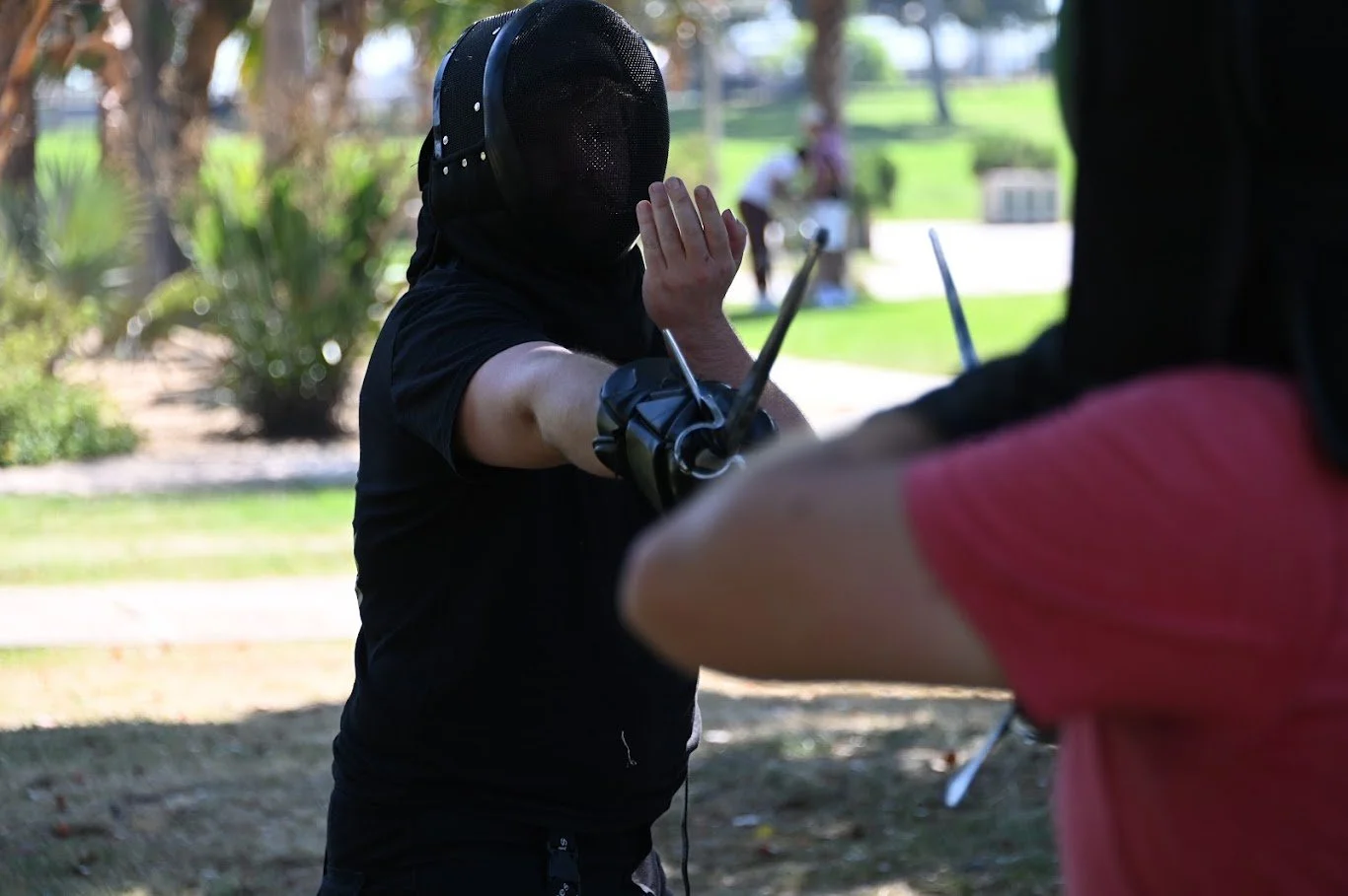 Person in fencing gear practicing with a fencing sword outdoors, with a person in a pink shirt partially visible in the foreground.